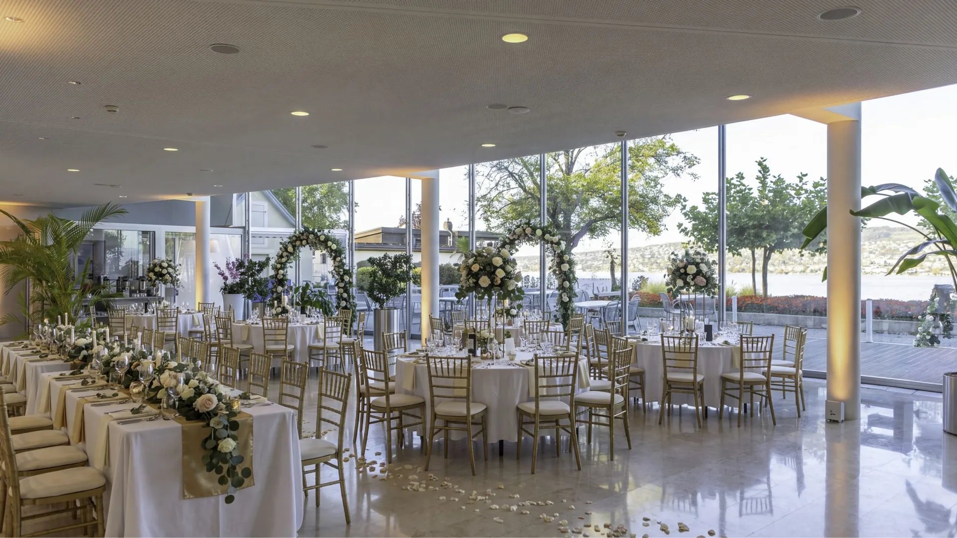 Lake-view event hall at LAGO Horgen with floor-to-ceiling windows onto Lake Zurich, set with round tables, a long table and flower arches