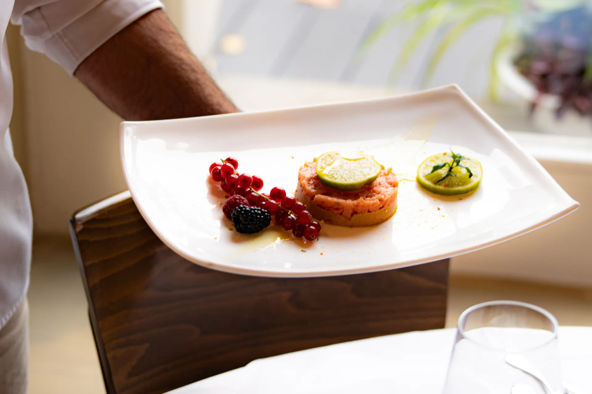 Ein Kellner bringt Lachs-Tatar mit Limette und frischen Beeren an einen Tisch am Fenster